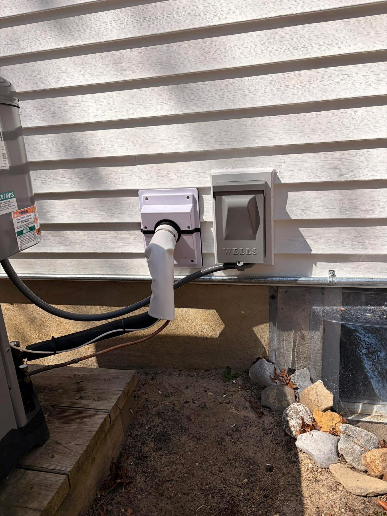 Outdoor electrical outlet on a metal wall with cables connected, surrounded by rocks and wooden steps.